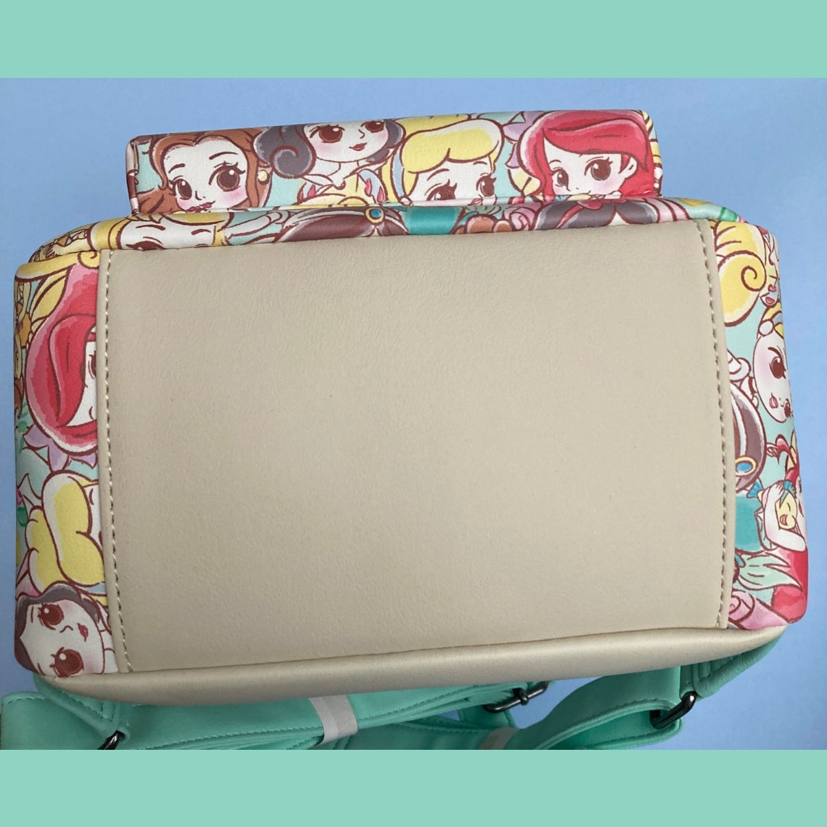 A close-up view of the pristine (large beige faux-leather panel) bottom of a NWT Chibi AOP Disney Loungefly mini backpack. Visible along the edges are princesses Ariel, Belle, Cinderella, and Snow White. Photo is trimmed by teal borders.
