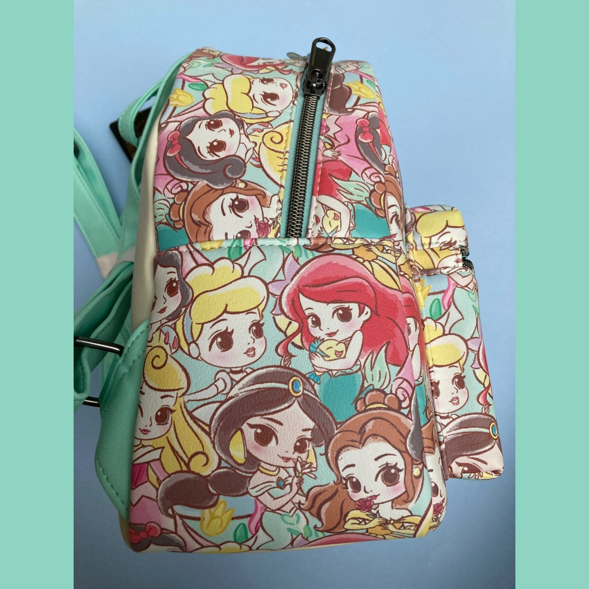 A Disney Loungefly mini backpack alternate side view featuring an all-over colorful Chibi princess print with teal faux-leather straps and trim, shown against a light blue background with teal side borders.