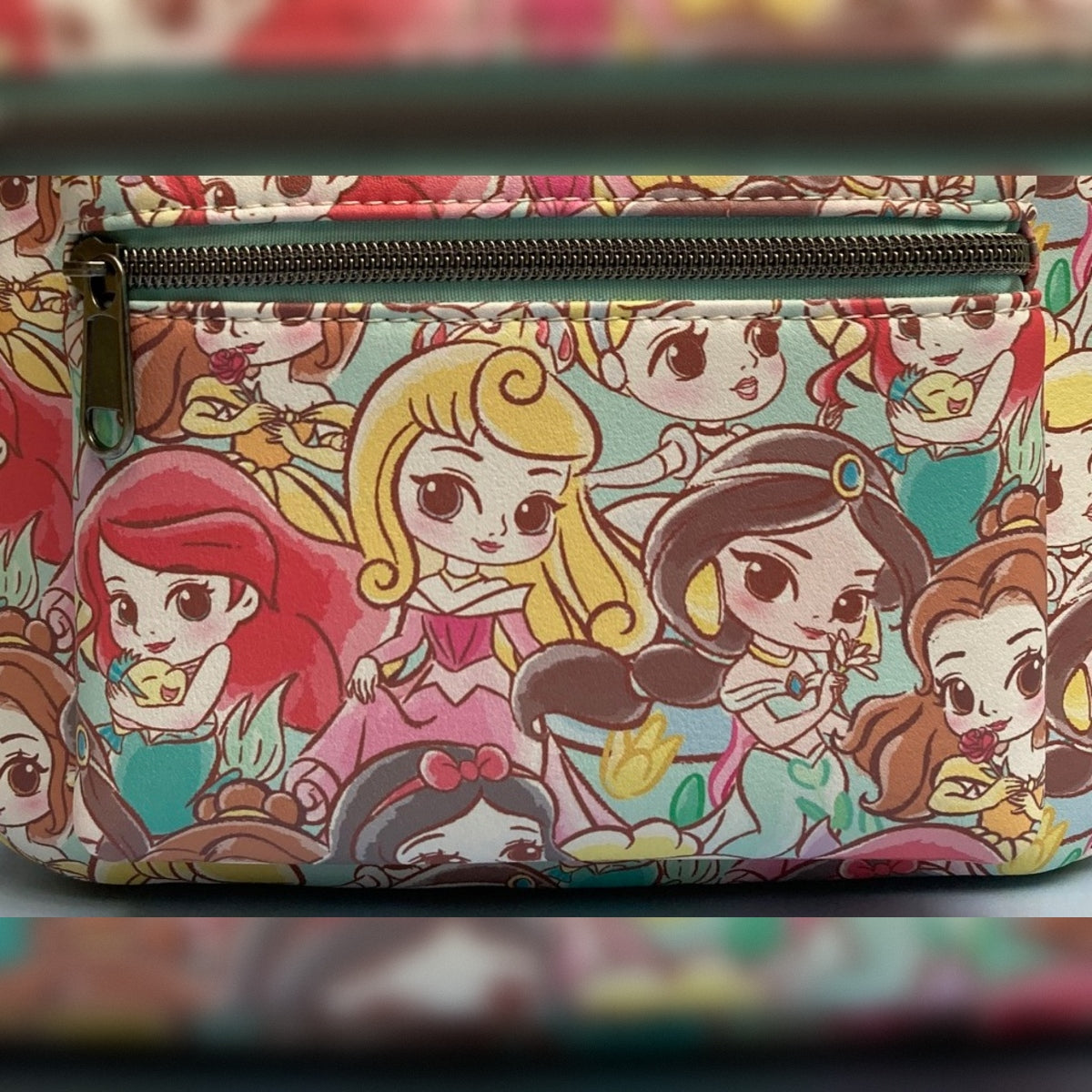 A close-up of the zippered front pocket of a Chibi AOP Disney Loungefly mini backpack featuring prime placement of Ariel, Aurora, Jasmine, and Belle nestled among other Disney Princesses on a pastel background.