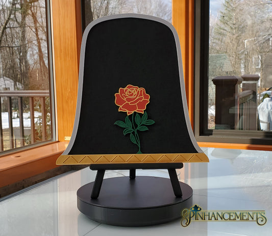 A black felt pinboard shaped like a bell jar with a silver arched border and a gold zigzag-patterned base, featuring a dimensional red and yellow rose with a green stem in the center. The pinboard is displayed upright on a small black easel, sitting on a glass table in a sunroom. The Pinhancements logo is in the bottom right corner.
