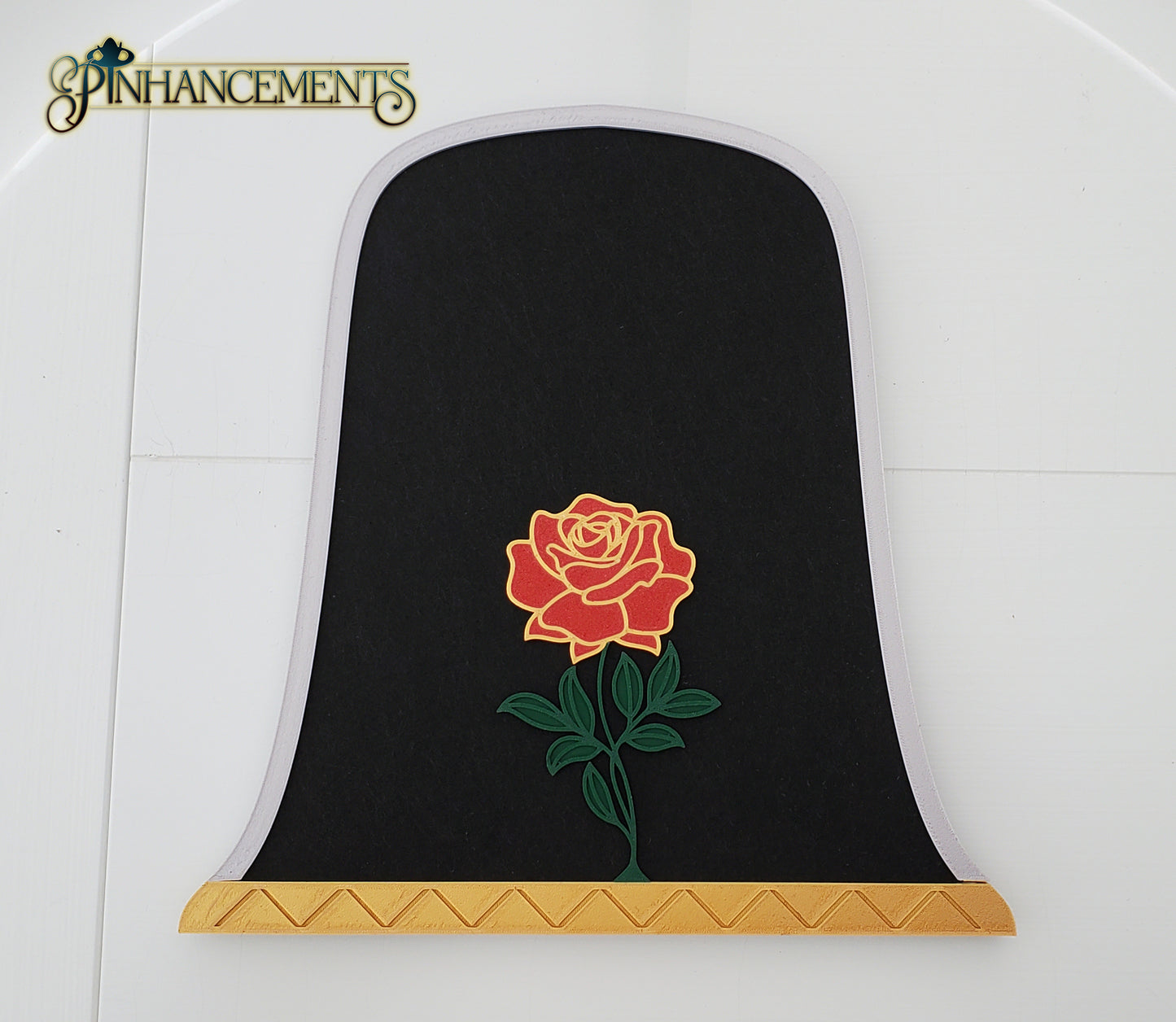A close-up of a bell jar-shaped black felt pinboard with a silver arched border, a gold patterned base, and a prominent 3D red rose with green leaves in the center. The pinboard is laid flat against a white paneled background. The Pinhancements logo is in the top left corner.