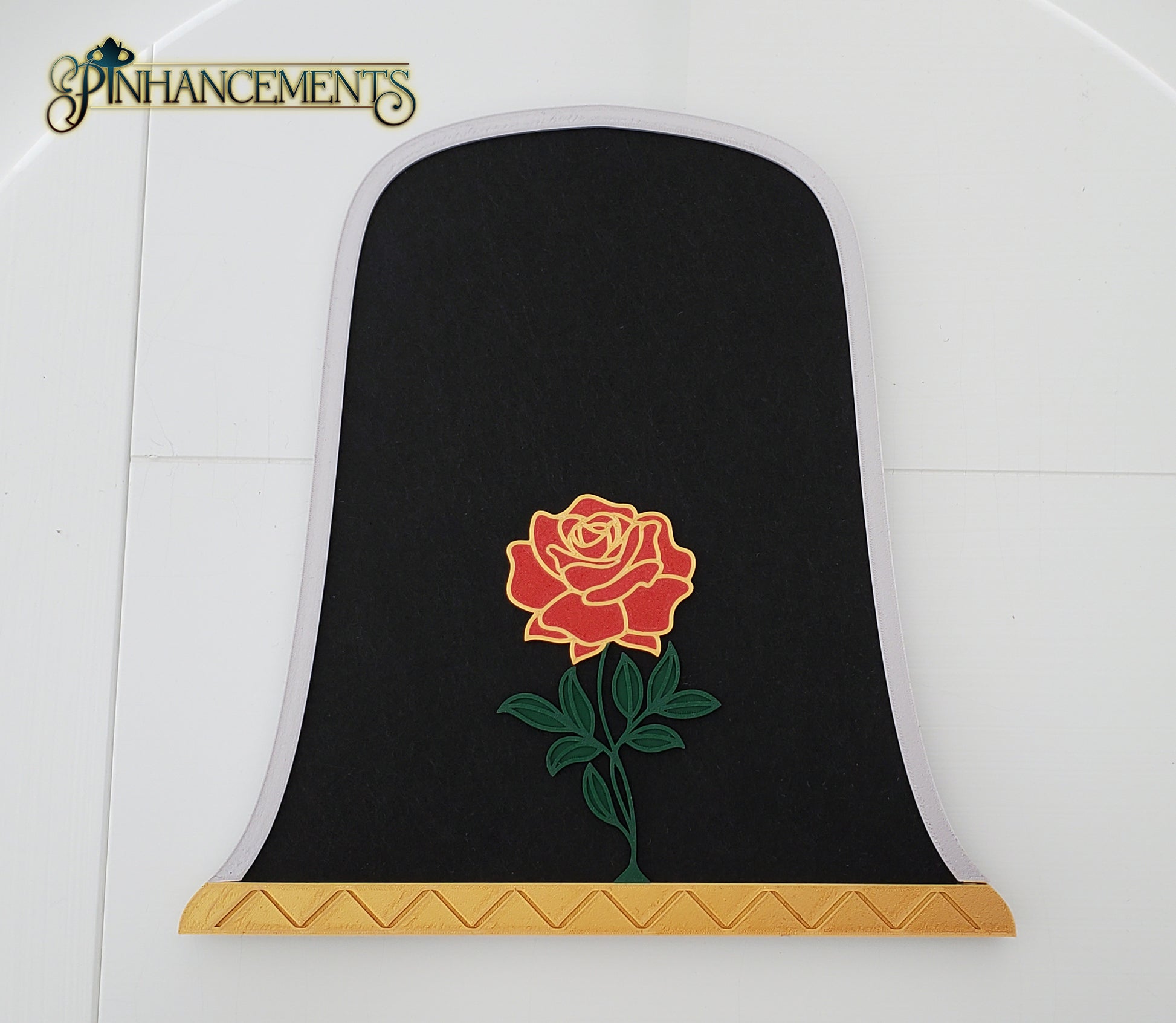 A close-up of a bell jar-shaped black felt pinboard with a silver arched border, a gold patterned base, and a prominent 3D red rose with green leaves in the center. The pinboard is laid flat against a white paneled background. The Pinhancements logo is in the top left corner.