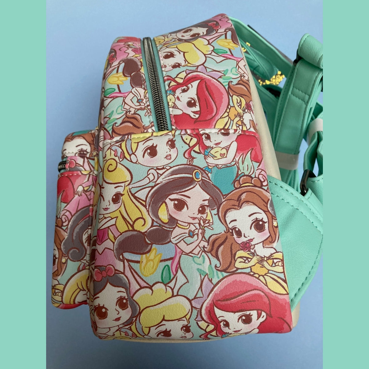 A Disney Loungefly mini backpack side view featuring an all-over colorful Chibi princess print with teal faux-leather straps and trim, shown against a light blue background with teal side borders.