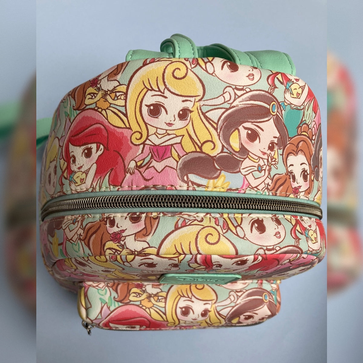 A Disney Loungefly mini backpack featuring an all-over colorful Chibi princess print, with a green faux-leather Disney Loungefly logo patch on the front and a zippered front pocket, photographed against a soft blue-gray blurred background.