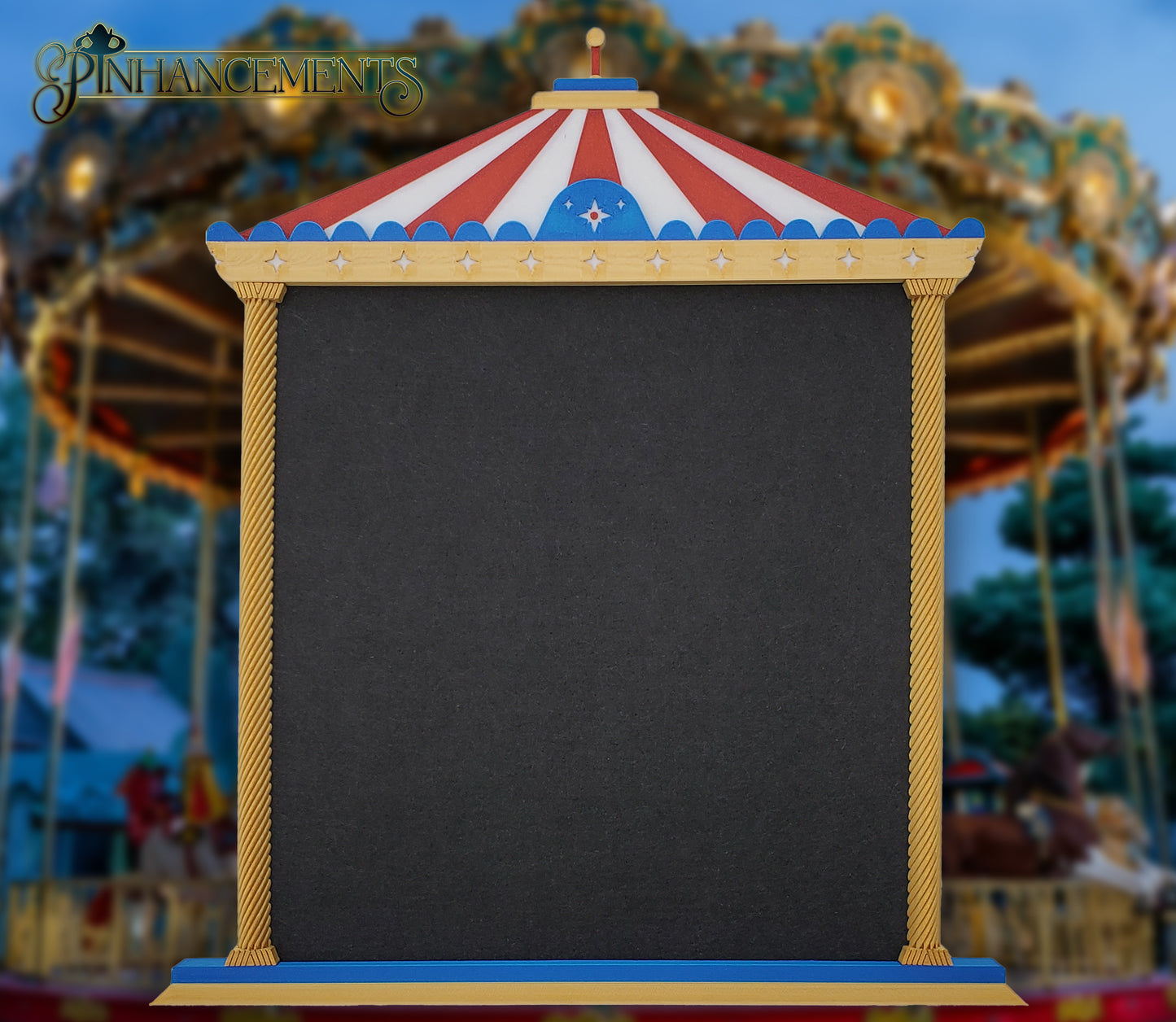 A straight-on view of a Pinhancements 3D-printed, carousel-themed pinboard. The frame features a red and white striped canopy, blue trim with star details, and twisted gold pillars bordering a large, blank black felt center for displaying enamel pins.