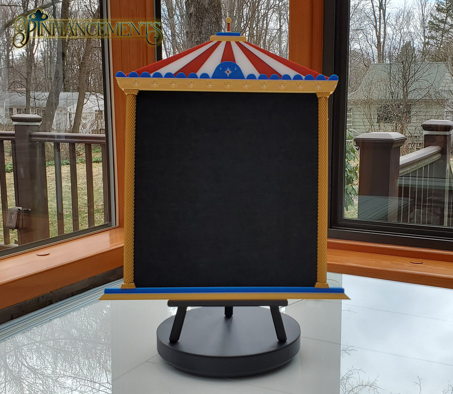 Gemini said
A Pinhancements carousel-themed pinboard displayed on a black tripod easel. The 3D-printed frame features a red and white striped canopy, blue trim with stars, and twisted gold pillars surrounding a blank, black felt center designed for displaying enamel pins