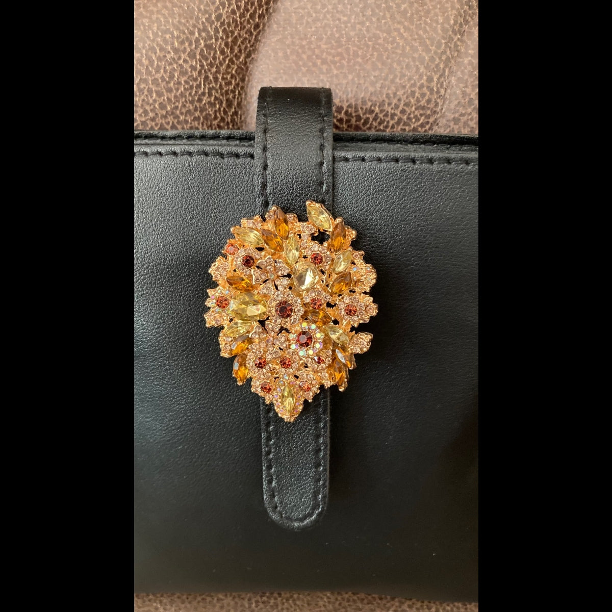 A close-up of a black rhinestone crossbody purse styled on a chair featuring a decorative gold-tone floral clasp embellished with multiple colored gemstones in yellow, orange, and red tones.