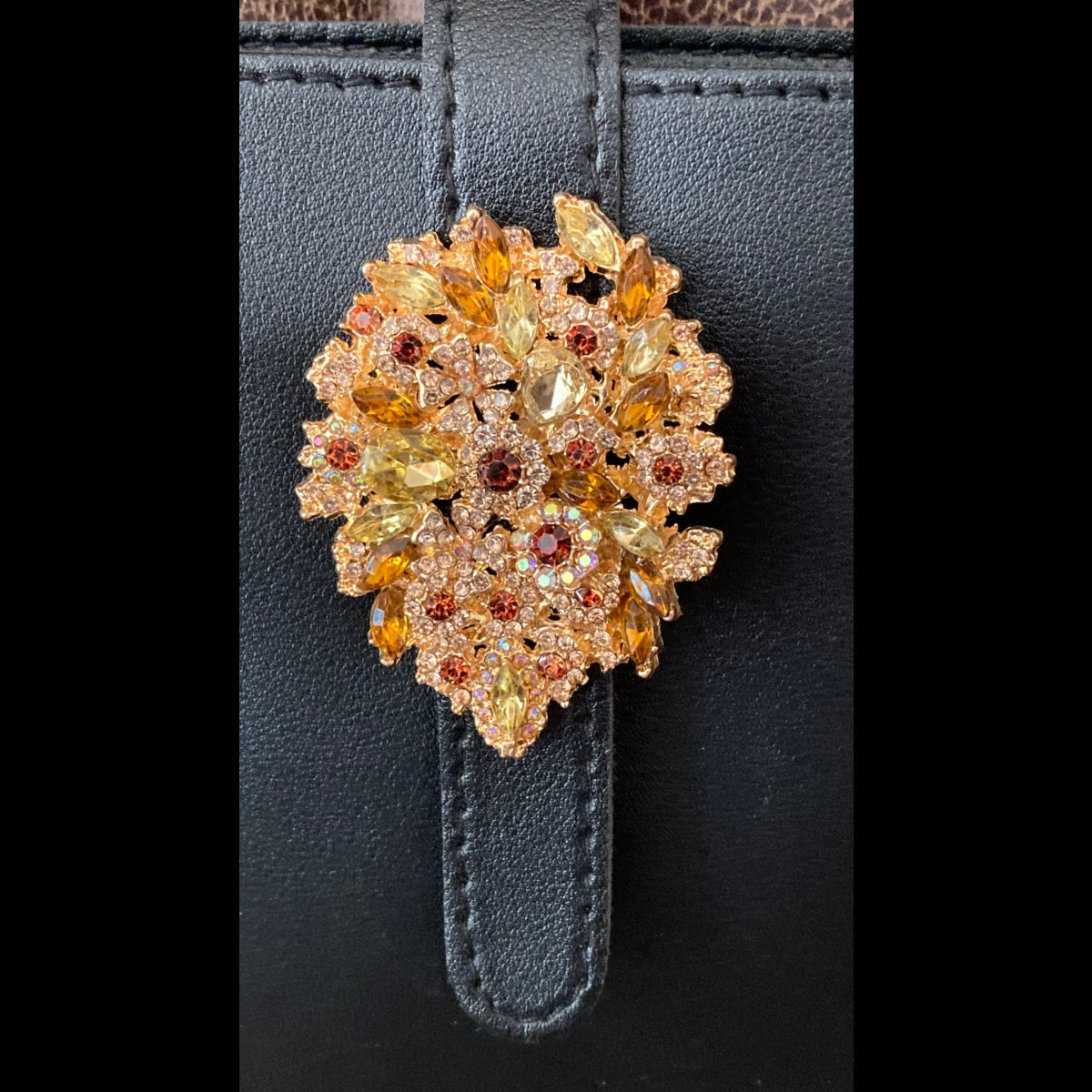 Black rhinestone crossbody purse styled on a chair, alternate angle featuring a close-up of a circular gold-toned brooch  with faceted amber, yellow, and reddish stones, attached to a black strap.