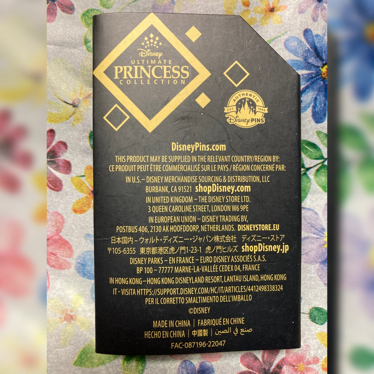 A black Disney Ultimate Princess Collection product tag (for Tiana) with gold text is photographed flat against a floral background. Visible Disney branding appears prominently on the tag.
