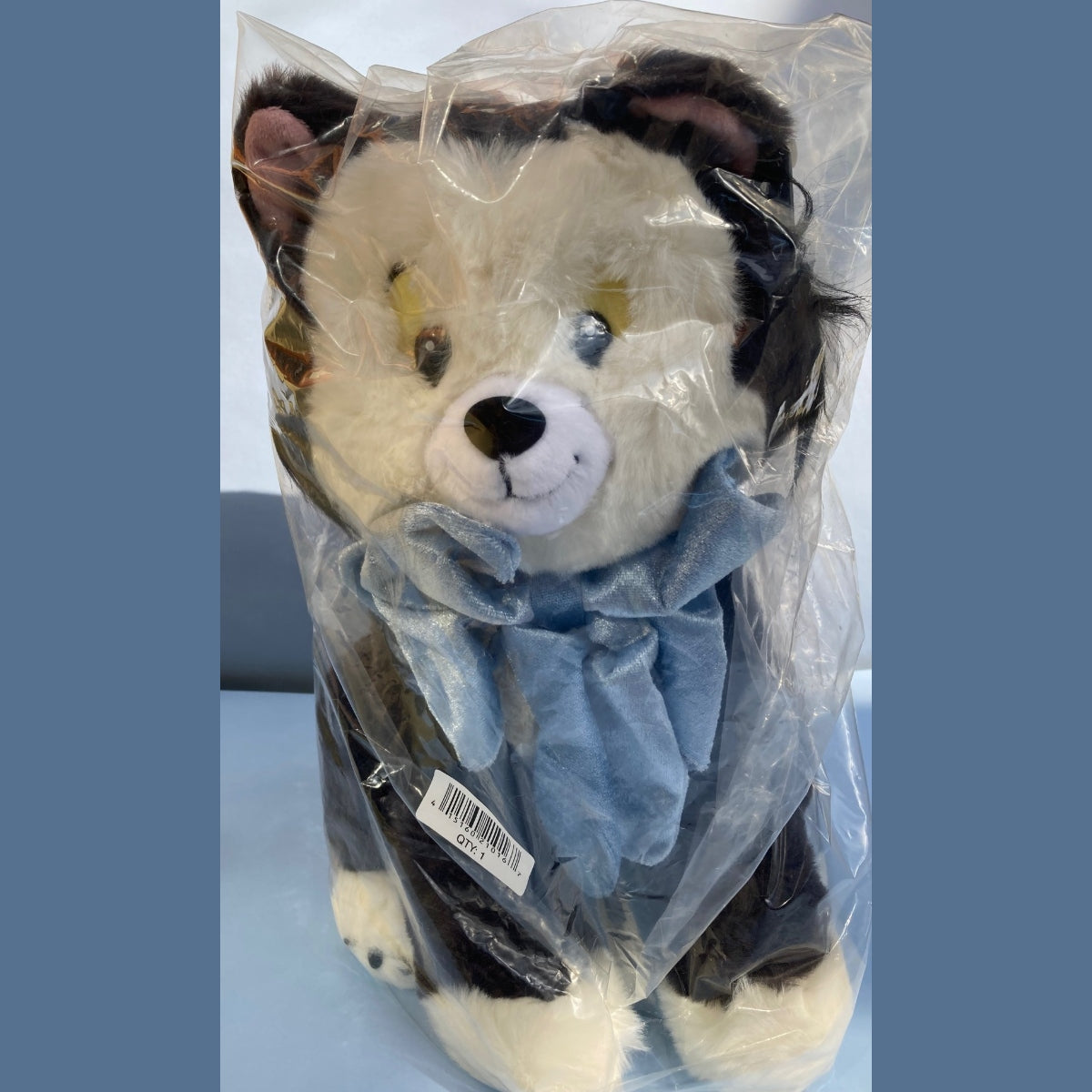 A Disney Figaro Plush factory wrapped in clear plastic packaging. Figaro has a cream-colored face and paws, black ears and a light blue scarf.