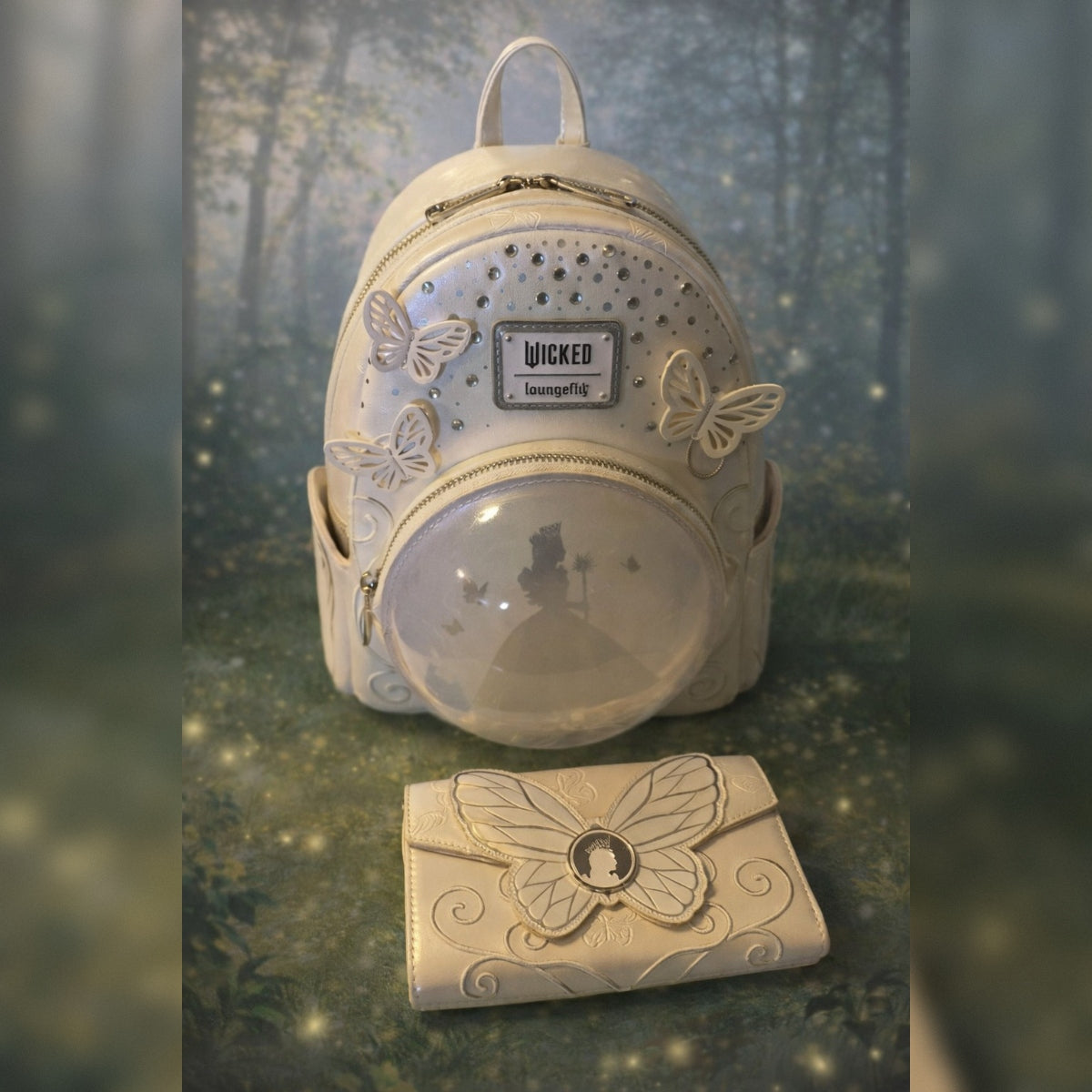 A white mini backpack with a clear bubble window showing Glinda with her wand, accompanied by a matching white wallet with a butterfly design. The backpack features rhinestone embellishments, a silver 'Wicked' logo patch, and 3D butterfly accents, set against a blurred forest background.