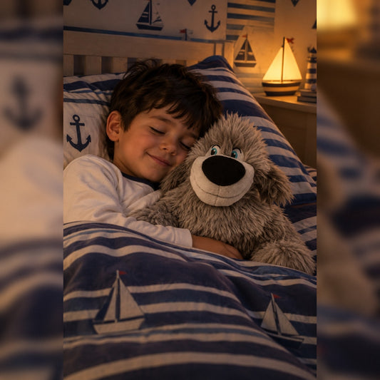 A young boy with dark hair lies in bed under a blue and white striped duvet with sailboat prints, cuddling a Disney Max plush dog. The room has a warm, cozy ambiance with a lit lamp on the bedside table.