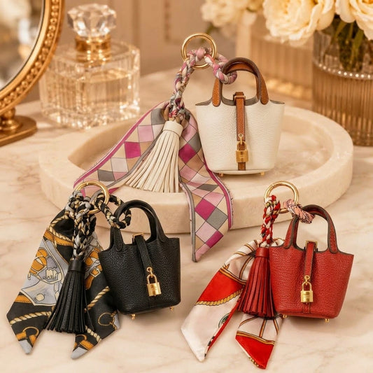 Three mini handbag-shaped keychains with leather-look bodies, gold-tone padlock details, and hanging scarves are arranged on a marble surface. The bags come in black, white, and red, each styled with a different patterned scarf.