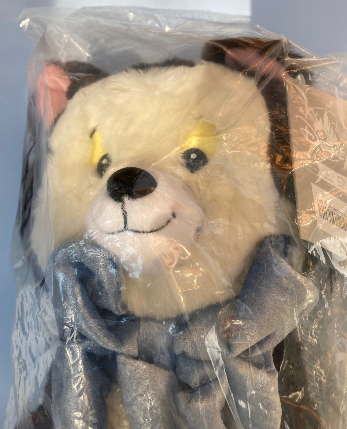 A Disney Figaro plush face wrapped in clear plastic packaging, with cream-colored fur, a black nose, and a blue scarf.