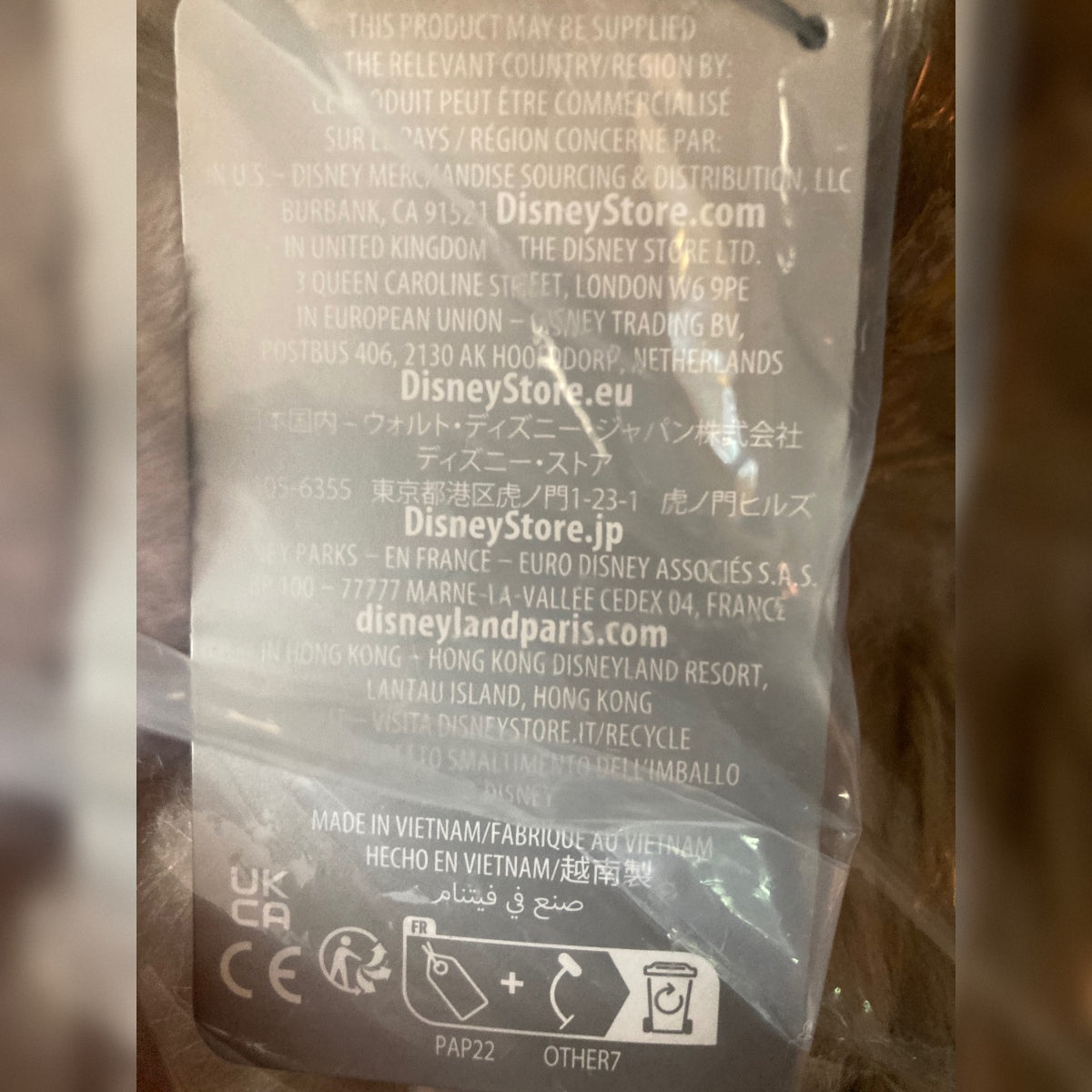 Close-up of the back of a Disney-branded tag in a packaging bag with printed distribution and website text. Photographed in a real environment with some glare and plastic reflections.