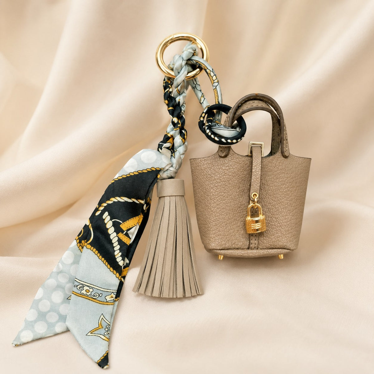 A miniature taupe pebbled handbag keychain charm with a matching tassel and braided scarf keychain, displayed on draped beige fabric.