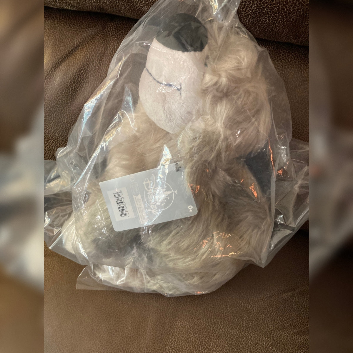 A Disney Max plush toy wrapped in original packaging is resting on a brown leather chair. The toy has a fluffy cream and brown fur-like texture, a white face and a black nose. A grey Disney hang tag is attached to the toy.