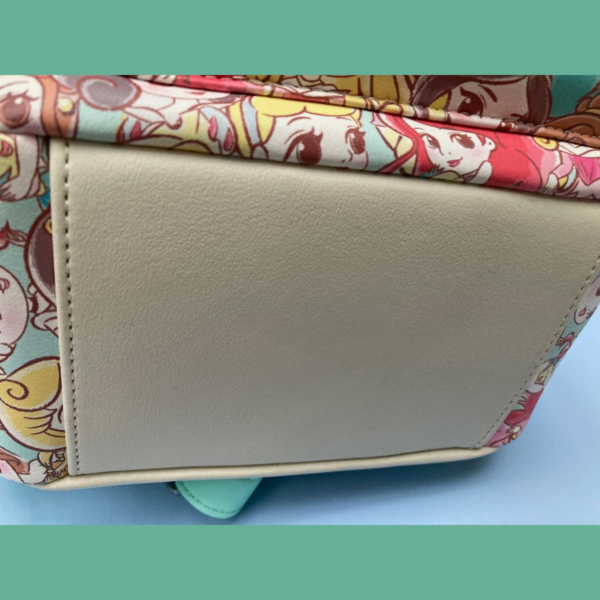 A close-up view of the pristine (large beige faux-leather panel) bottom of a NWT Chibi AOP Disney Loungefly mini backpack. Visible along the edges are princesses Ariel, Jasmine, and Cinderella.