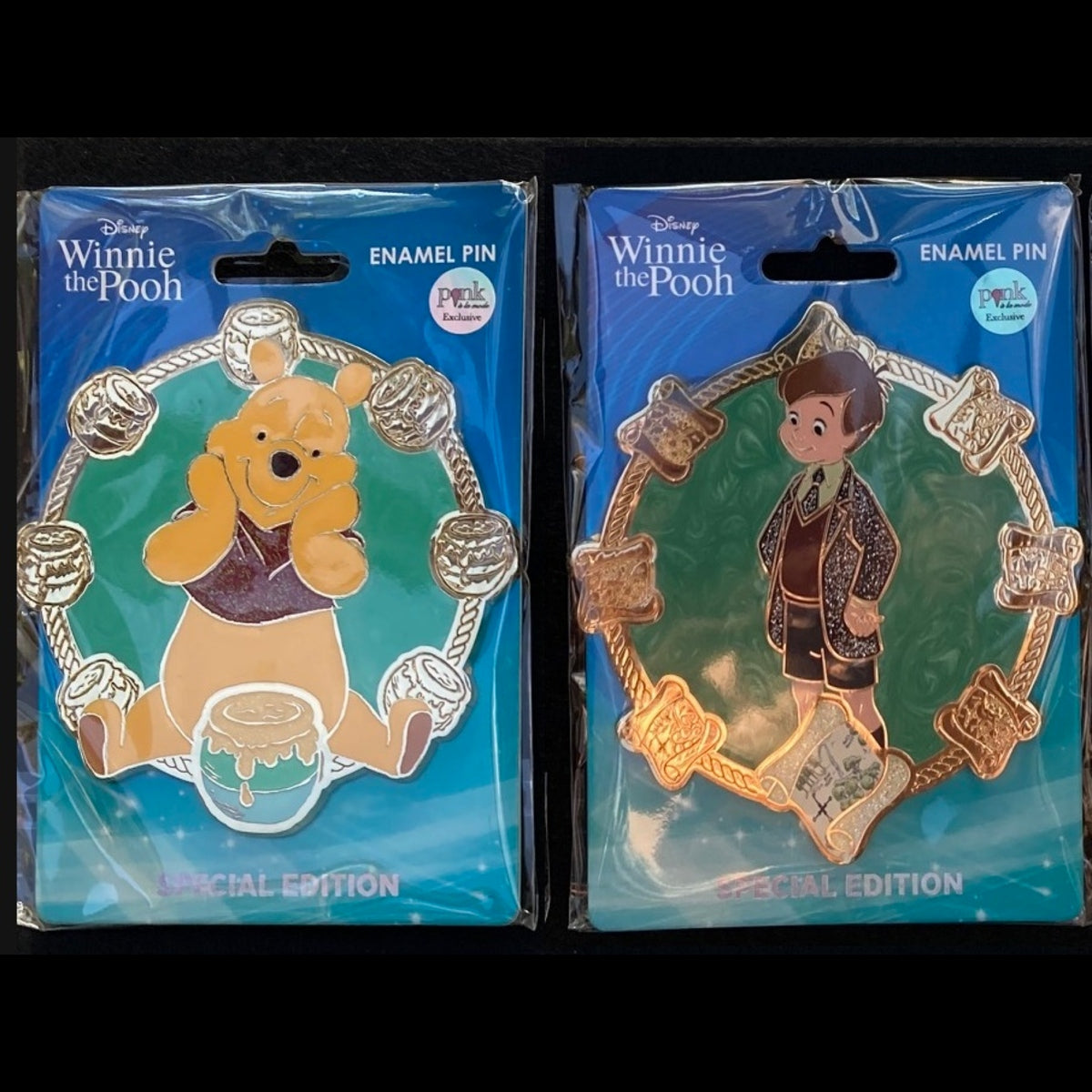 Two Disney Winnie the Pooh enamel pins in original blue packaging, shown side by side on a black background. The left pin features Pooh holding a honey pot, and the right pin features Christopher Robin with maps.