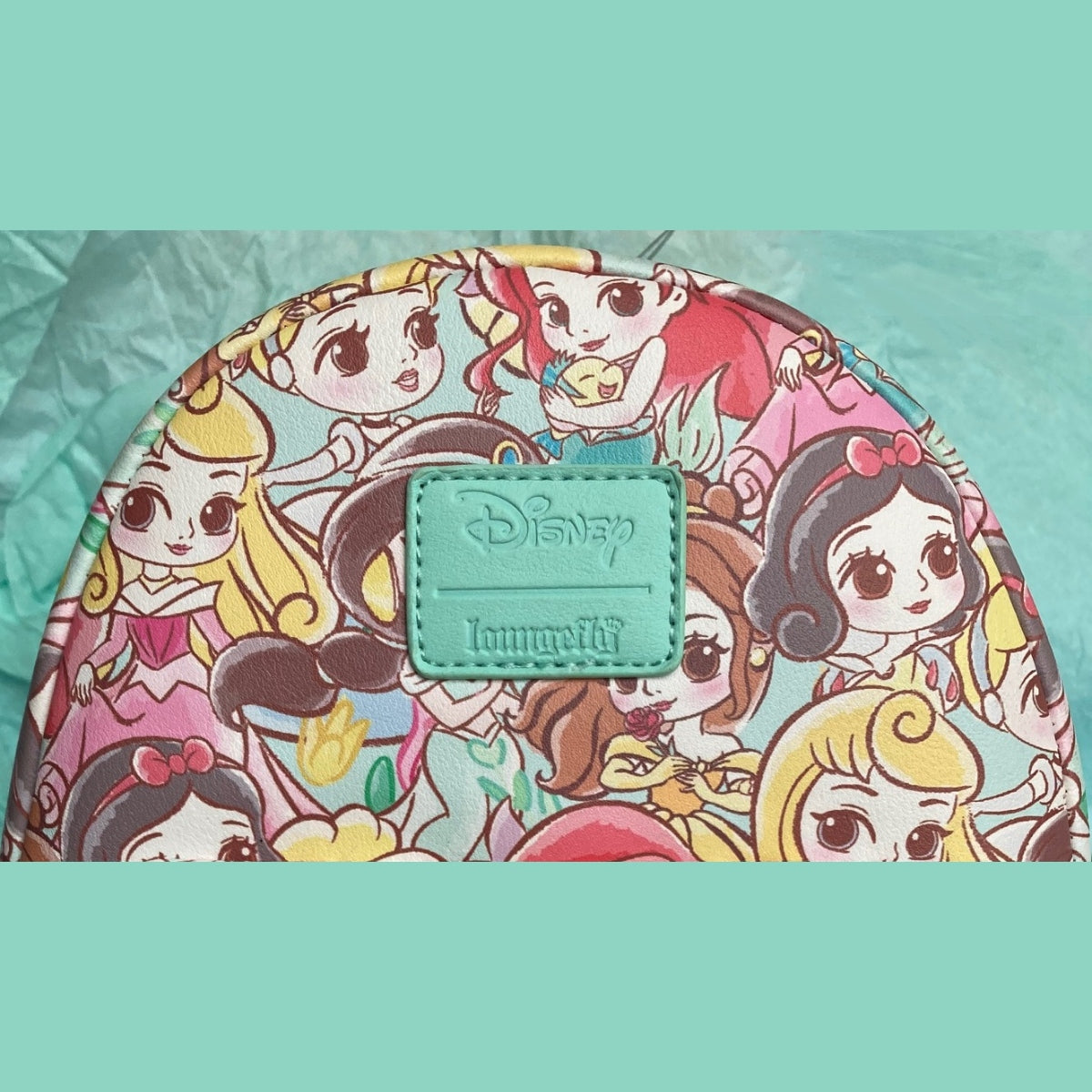 A close-up view of the top half of a Chibi AOP Disney Loungefly mini backpack featuring Aurora Cinderella, Ariel, and Snow White, surrounded by teal borders.