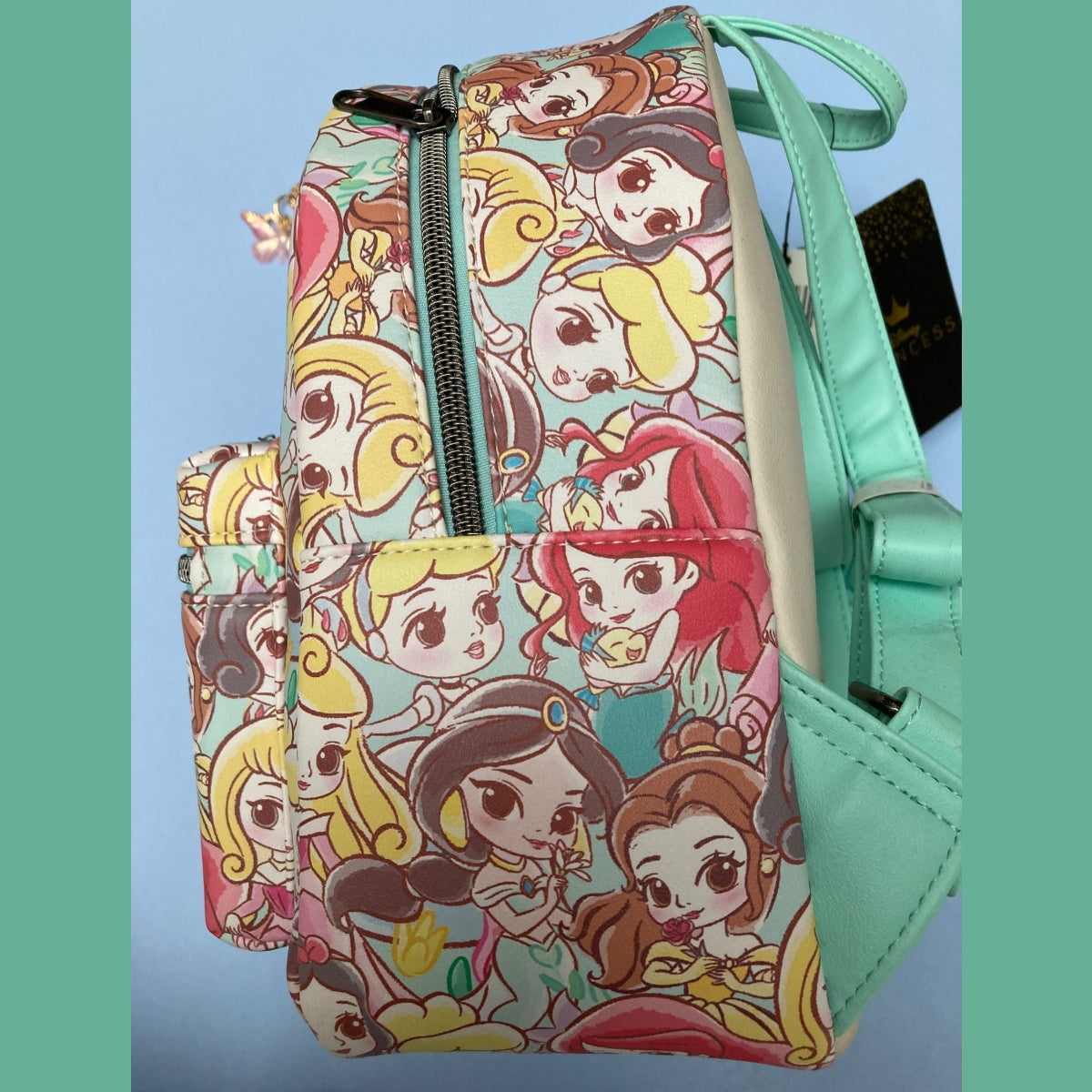 A Disney Loungefly mini backpack side view featuring an all-over colorful Chibi princess print with teal faux-leather straps and trim, shown against a light blue background with teal side borders.