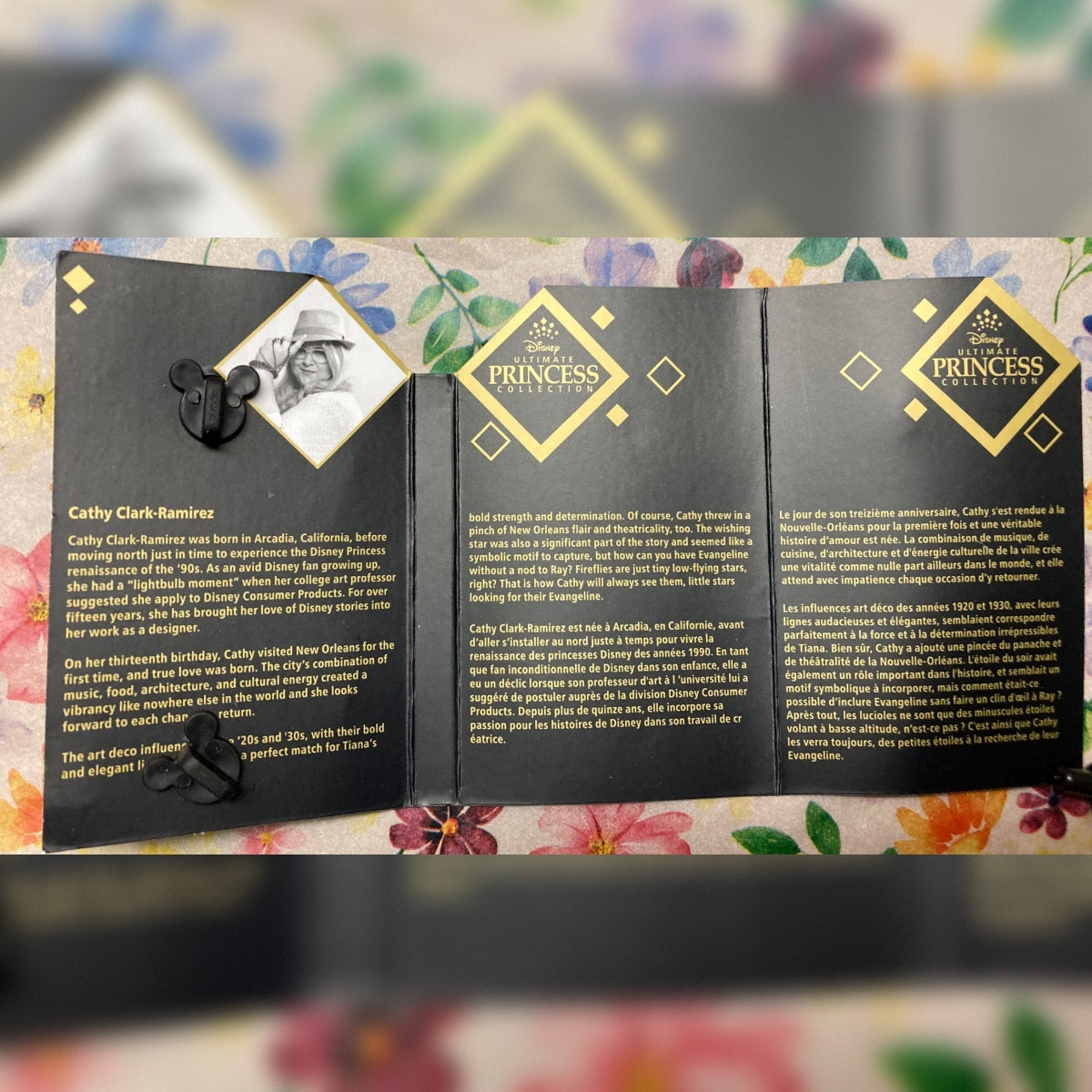 A three-panel black Product Tag featuring a portrait of Cathy Clark Ramier, French and English text, and gold Disney Ultimate Princess Collection logo, displayed on a floral surface.
