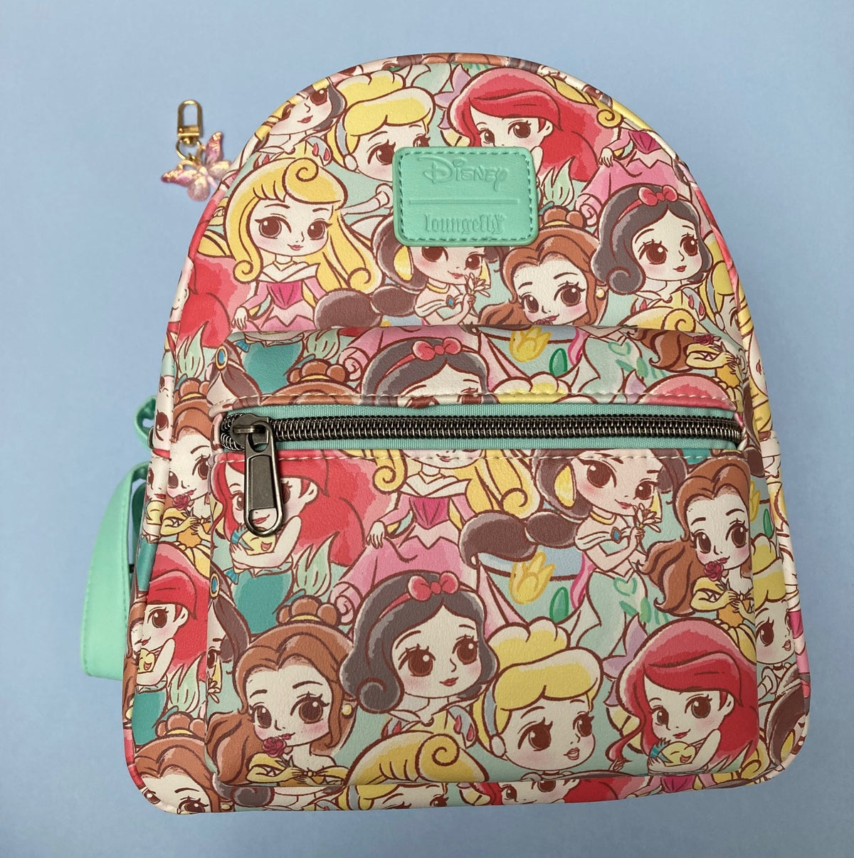 A mini Disney Loungefly backpack featuring an all-over colorful Chibi princess print, with a green faux-leather Disney Loungefly logo patch on the front and a zippered front pocket. Also included is a removable pink butterfly zipper pull