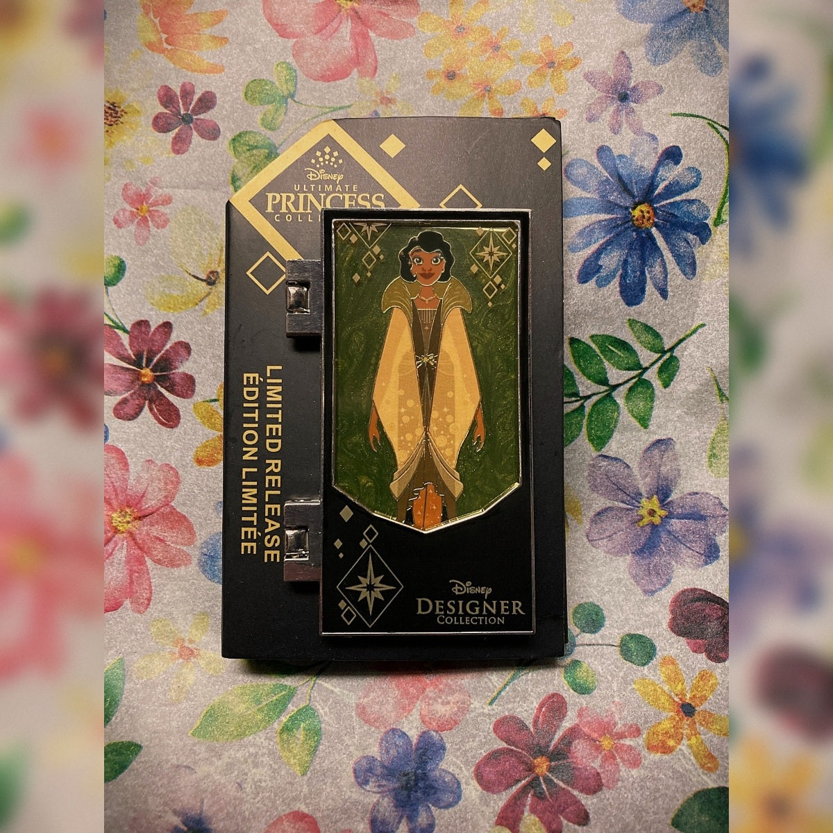 An image of the Tiana Pin from the Ultimate Princess Collection on its black backer card on top of a floral background.
