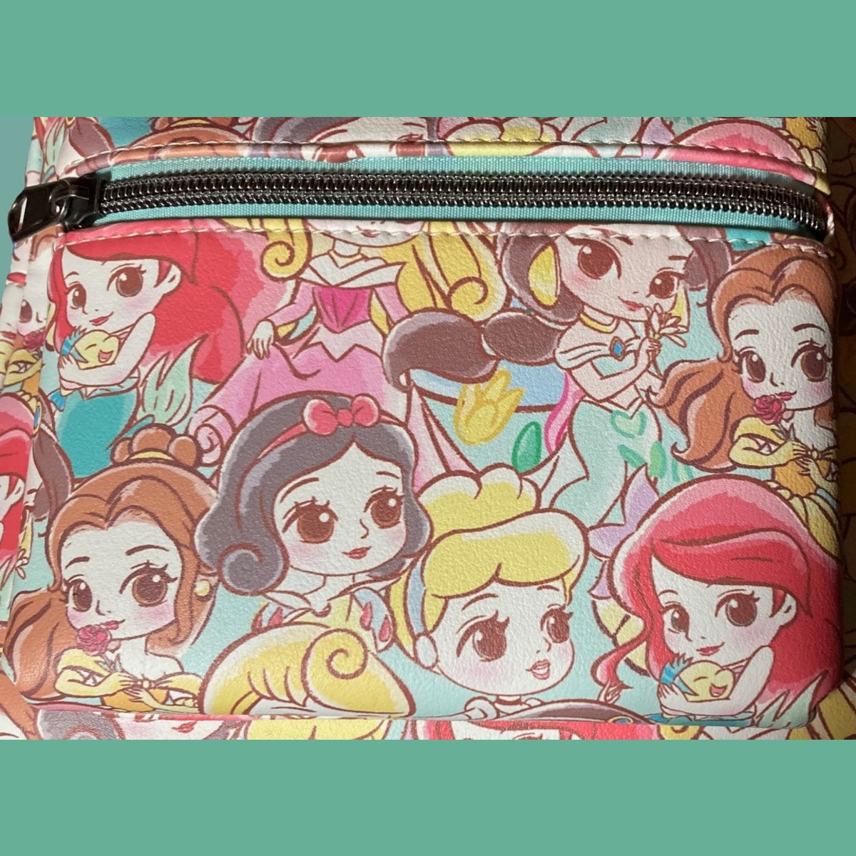 A close-up of the zippered front pocket of a Chibi AOP Disney Loungefly mini backpack featuring prime placement of Ariel, Belle, and Snow White, with Cinderella, Jasmine, and Belle nestled among other Disney Princesses on a Teal background.