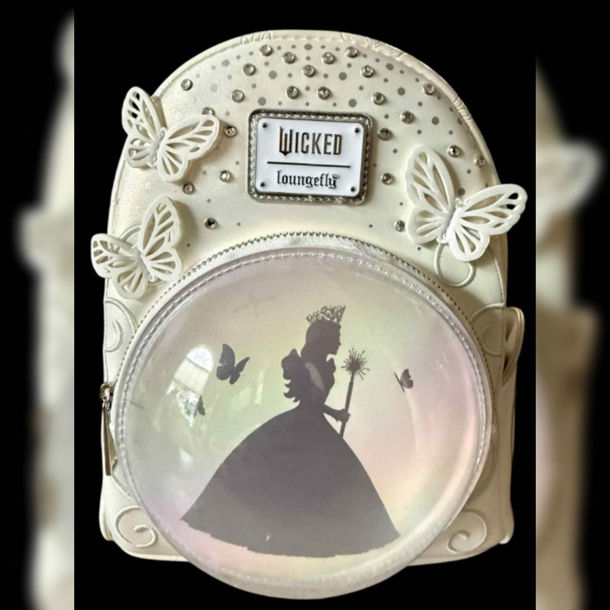 A decorative mini backpack featuring a cream-colored faux leather body adorned with white butterfly embellishments, rhinestone studs, and a central 'Wicked Loungefly' brand plaque. The front has a large clear oval window displaying a  silhouette of a silhouette of Glinda in a ballgown holding a wand, with small butterfly silhouettes inside.
