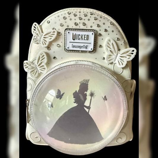 A decorative mini backpack featuring a cream-colored faux leather body adorned with white butterfly embellishments, rhinestone studs, and a central 'Wicked Loungefly' brand plaque. The front has a large clear oval window displaying a  silhouette of a silhouette of Glinda in a ballgown holding a wand, with small butterfly silhouettes inside.